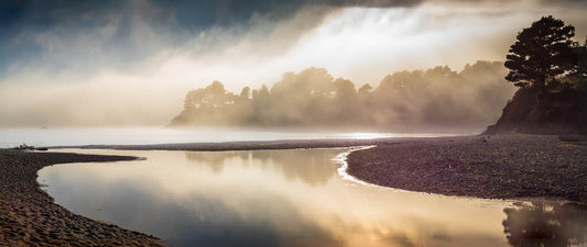 Photo of a brightly lit fog bank and an ocean bay, luminous, against a pale headland in the hazy distance.
